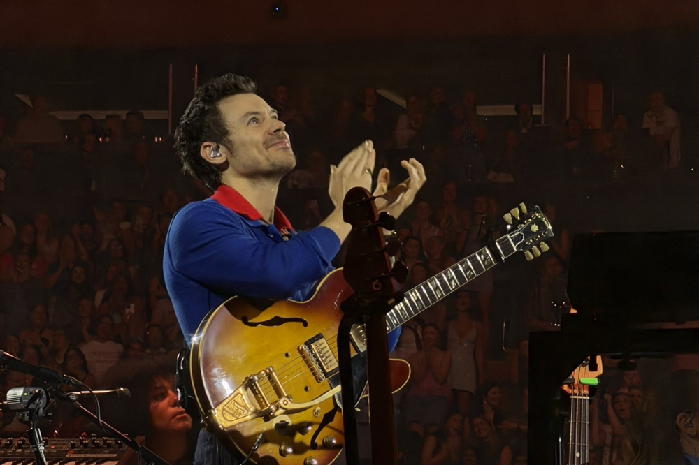 Harry Styles no palco, sorrindo e aplaudindo, usando camisa azul com gola vermelha e segurando uma guitarra semiacústica marrom. O público aparece desfocado ao fundo