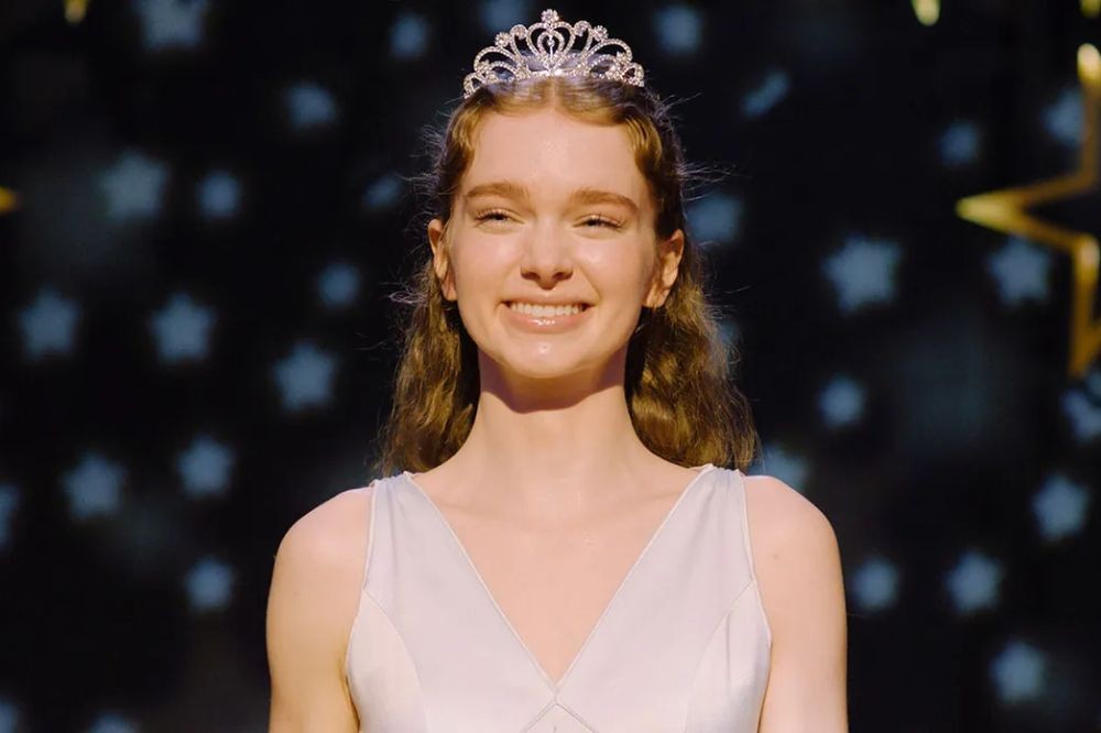 Jovem mulher de pele clara, cabelo castanho-claro ondulado e tiara prateada brilhante, sorrindo para a câmera. Veste um vestido branco de alças finas, com decote em V. O fundo escuro tem estrelas azuis e uma estrela dourada maior à direita