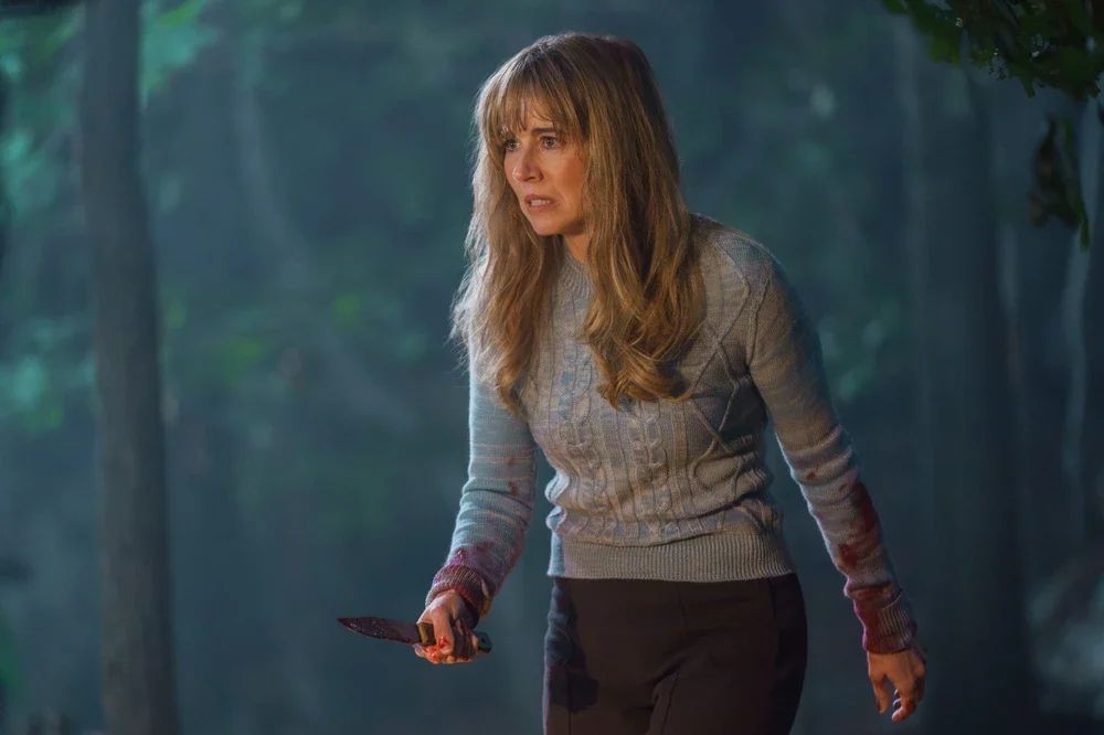 Mulher loira, com franja e cabelos longos, veste suéter azul claro com manchas de sangue nos punhos, segurando uma faca ensanguentada. Ela tem expressão assustada, olhando para a direita, em uma floresta escura com névoa