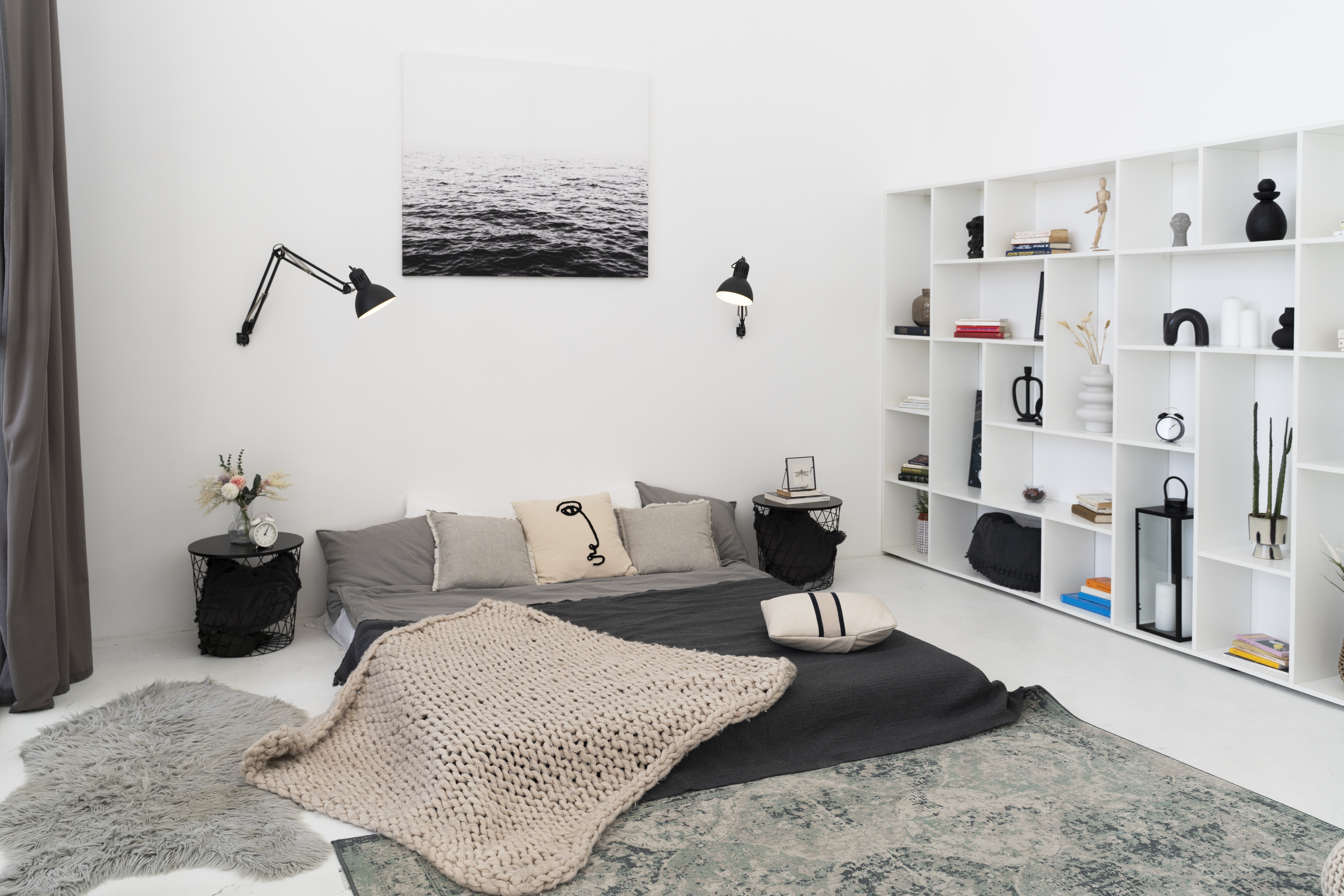 Quarto moderno com cama baixa cinza, almofadas claras e uma manta de tricô bege. Duas mesas de cabeceira pretas com flores e um relógio. Na parede, uma imagem em preto e branco do mar e duas luminárias articuladas. Uma estante branca cheia de objetos decorativos e livros. Tapetes cinzas no chão.