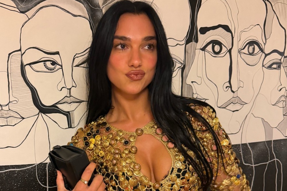 Dua Lipa, com longos cabelos pretos, usa um vestido dourado brilhante com decote e recortes, fazendo bico para a câmera. Ao fundo, uma parede com desenhos abstratos de rostos em preto e branco.