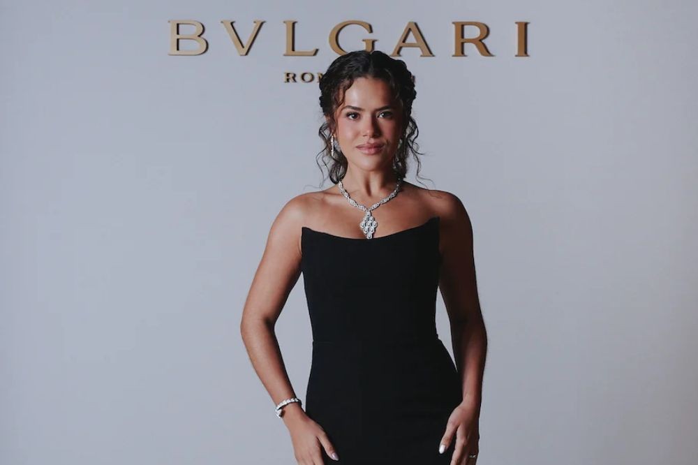 Mulher de pele morena e cabelos cacheados presos, sorrindo, usando um vestido tomara que caia preto, colar e pulseira de diamantes, posando em frente a uma parede clara com o logotipo dourado BVLGARI ROMA