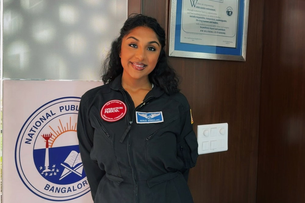 Mulher indiana de cabelo escuro e ondulado, sorrindo, vestindo um macacão preto com patches vermelhos e azuis, em frente a um logo circular branco e uma parede de madeira