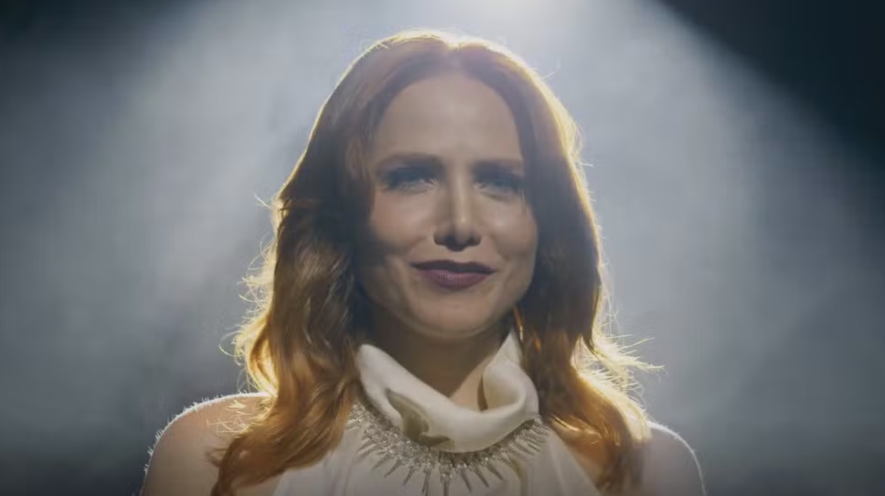 Mulher de cabelos ruivos e ondulados, sorrindo levemente, usa um vestido branco com gola alta e um colar prateado de pontas. Ela está sob um foco de luz forte, com fumaça ao fundo