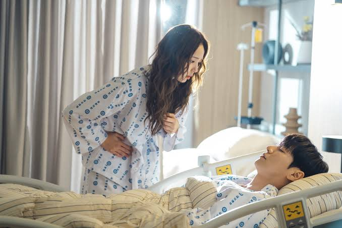 Uma mulher de cabelos longos e ondulados, vestindo pijama hospitalar branco com bolinhas azuis, inclina-se sobre um homem deitado em uma cama de hospital. Ela tem uma expressão preocupada e segura a barriga. O homem, também de pijama hospitalar, parece dormir ou estar inconsciente. O quarto tem cortinas claras e um suporte de soro ao fundo